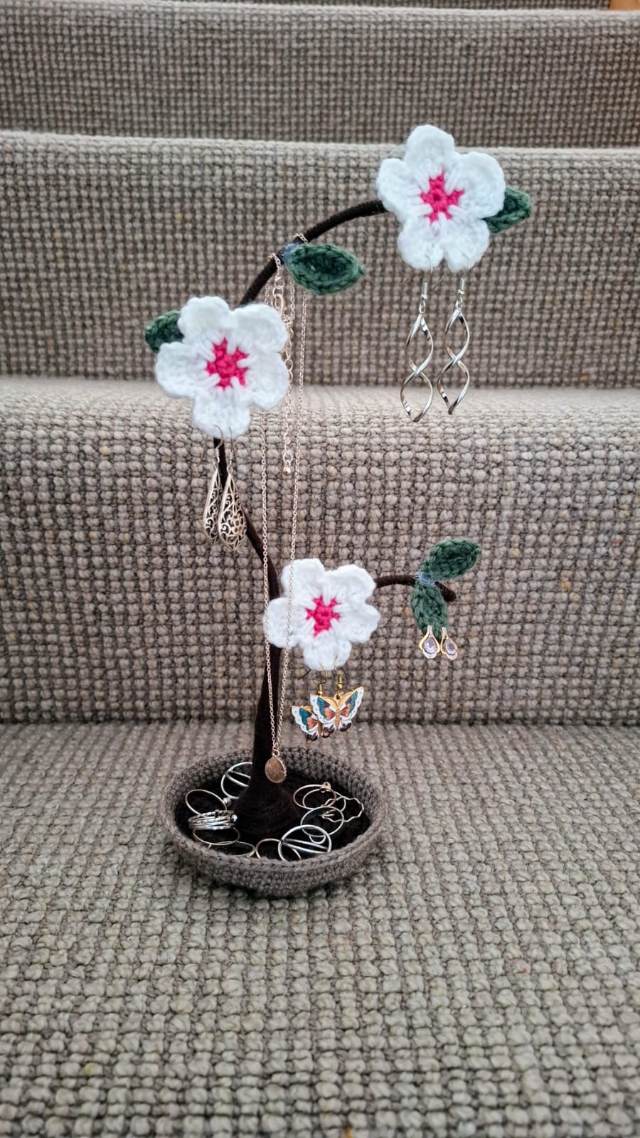 A crocheted jewellery tree with white cherry-blossom flowers holding earrings and necklaces, with a crochet dish base